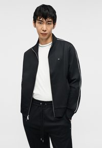 Μαύρο μπουφάν με φερμουάρ και λευκές ρίγες, φορεμένο πάνω από λευκό μπλουζάκι. Διαθέτει άνετη εφαρμογή και παντελόνι με ελαστική μέση.