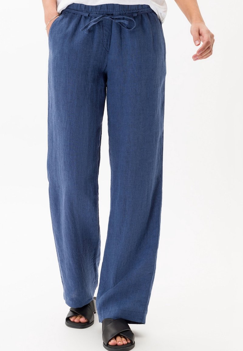 Personne portant un pantalon en lin bleu ample avec taille à cordon et des sandales noires ouvertes à l’avant sur fond blanc.