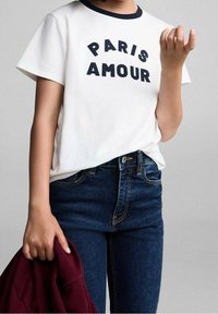 Camiseta de algodón blanca con cuello de canalé en azul marino, que presenta "PARIS AMOUR" en letras gruesas azul marino. Combinada con jeans de mezclilla azul oscuro.