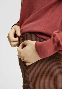 Bordowy longsleeve na brązowych spodni z kratą. Ręka zapina zamek w spodniach, które mają subtelny wzór w kratkę.