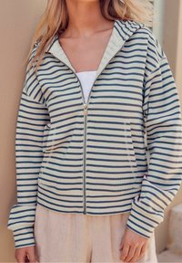Pruhovaná mikina na zip v světlé smetanové a námořnické barvě, s předním zipem, dvěma kapsami a kapucí; vyrobena z měkké tkaniny s volným střihem.