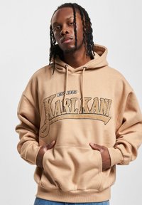 Bežinė sportinė palaidinė su priekiu kišene, turi juodą "KARL KANI" logotipą ir "EST 1989" užrašą. Medžiaga atrodo minkšta, su laisva siluetu ir su raišteliais.