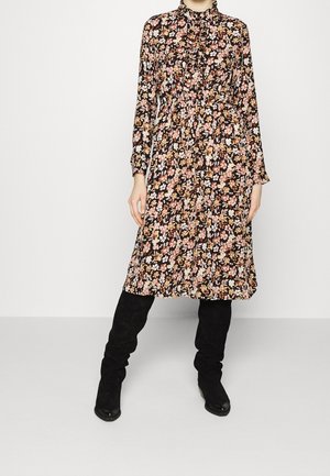 Femme portant une robe noire à manches longues avec un imprimé floral rose, orange et blanc, assortie à des bottes noires montantes, debout sur un sol gris.