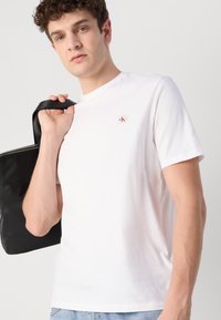 Camiseta blanca de algodón con cuello redondo, mangas cortas y un pequeño parche de logo. La persona sostiene una bolsa negra con cremallera.