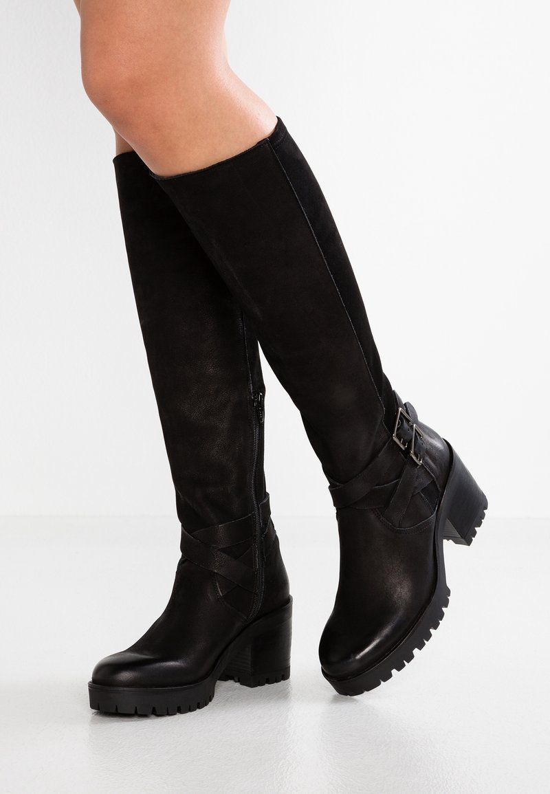 Piernas con botas de cuero negras hasta la rodilla, de tacón grueso y hebillas decorativas, de pie sobre un suelo blanco contra una pared blanca.