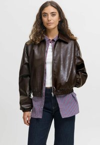 Hnědá kožená bomber bunda s elastickými manžetami a pasem, v kombinaci s modro-červenou kostkovanou košilí. Model stojí na neutrálním pozadí.