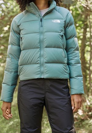 Persoon die een turquoise gewatteerd jack met het The North Face-logo en zwarte outdoorbroek draagt, staand in een bosrijke omgeving.