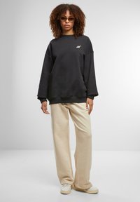 Svart oversized sweatshirt med rund hals og brodert logo, kombinert med kremfargede vide bukser og kremfargede sneakers. Krøllete hår og solbriller.