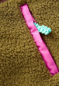 Bruine gestructureerde fleece stof met een roze binnenvoering die zichtbaar is in een naad, en een turquoise trekband voor aanpassing.