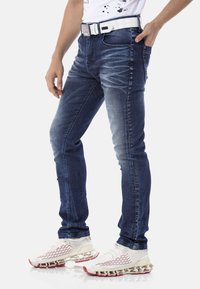 Jean en denim bleu avec une coupe slim, présentant une légère usure et des détails de froissage subtils. Associé à une ceinture blanche et des chaussures de sport.