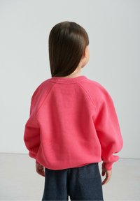 Różowa bawełniana bluza o luźnym kroju z rękawami raglanowymi, zestawiona z ciemnogranatowymi spodniami z polaru. Tkanina wydaje się miękka i o fakturze.