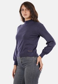 Maglione lavorato a maglia blu navy con colletto alto, silhouette aderente e maniche a sbuffo. Presenta un logo metallico "YES" nella parte anteriore in basso a sinistra.