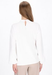 Blusa bianca a maniche lunghe con apertura a chiave sulla schiena, texture morbida, vestibilità rilassata e polsini elastici; abbinata a pantaloni beige.