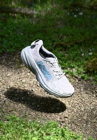Scarpa da corsa grigia con parte superiore in mesh traspirante, suola testurizzata per una migliore aderenza e branding HOKA blu. Design elevato con dettagli ammortizzanti.