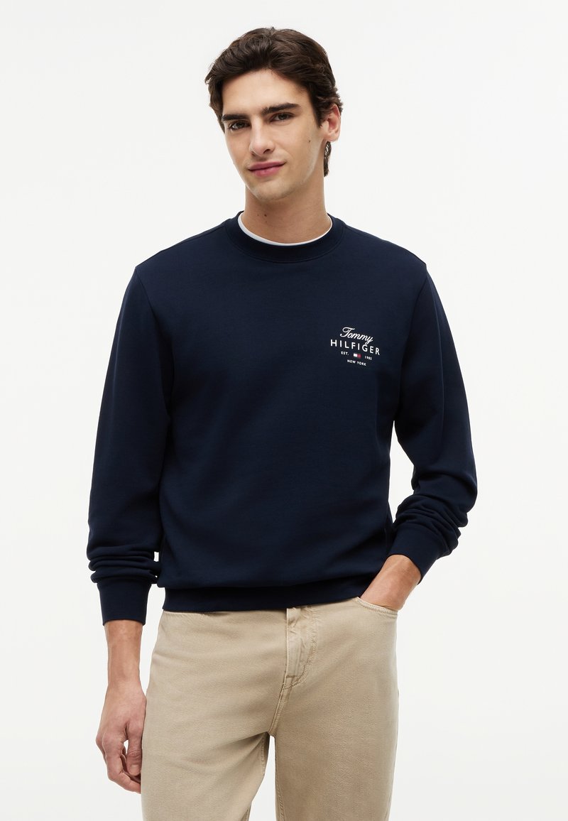 Jeune homme portant un sweat-shirt bleu marine avec le logo "Tommy Hilfiger" et un pantalon beige, debout une main dans la poche sur fond blanc.