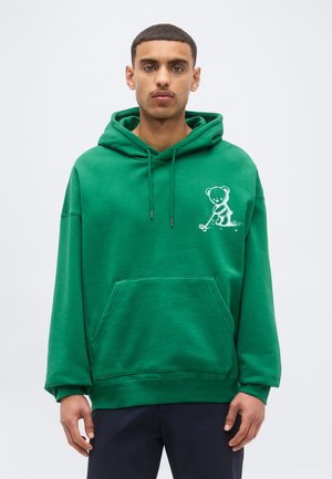 Młody mężczyzna w zielonej bluzie z kapturem z białym haftowanym na piersi misiem grającym w golfa, stoi na jednolitym tle.