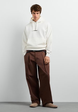 Jonge man met een witte hoodie, bruine wijde broekspijpen en beige schoenen, staand tegen een effen lichte achtergrond.