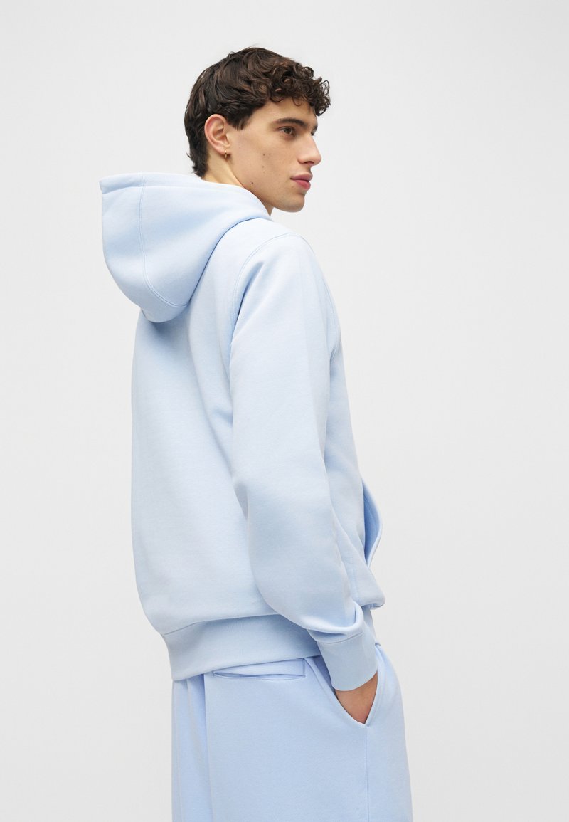 Joven con cabello rizado oscuro que lleva una sudadera con capucha azul claro y pantalones a juego, de pie con la mano en el bolsillo, mirando de lado.