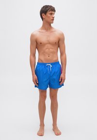 Jeune homme debout, pieds nus, torse nu, portant un short de bain bleu vif avec un cordon blanc, regardant vers sa gauche sur un fond uni.