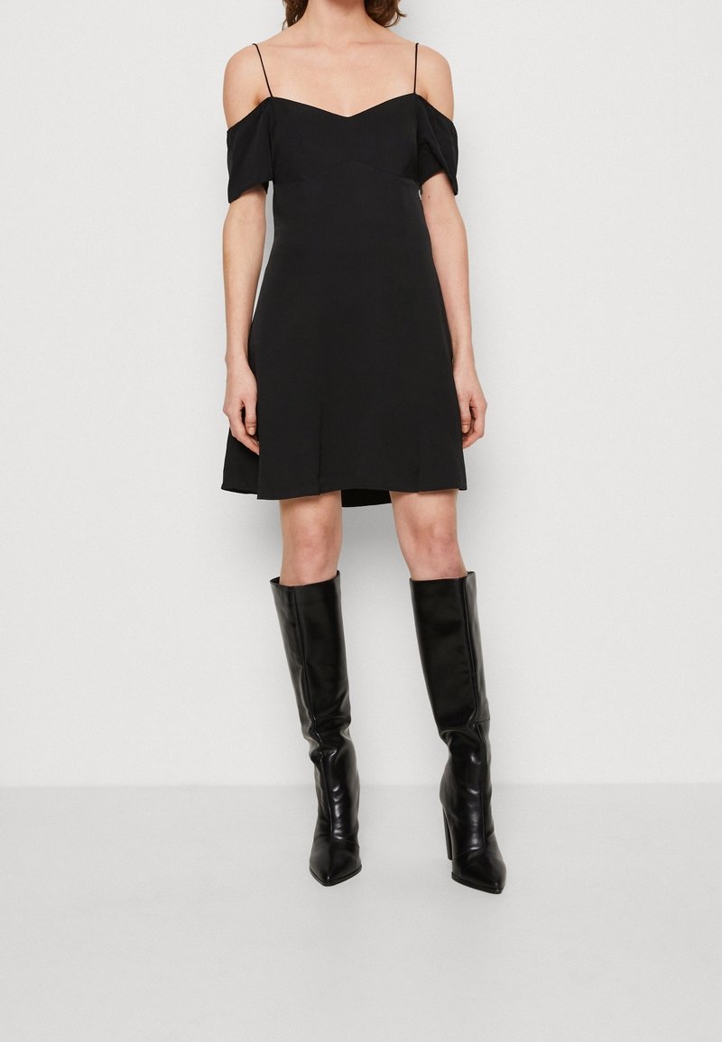 Robe noire à épaules dénudées avec un décolleté en V et une jupe courte évasée. Associée à des bottes noires en cuir à talons aiguilles jusqu'aux genoux.