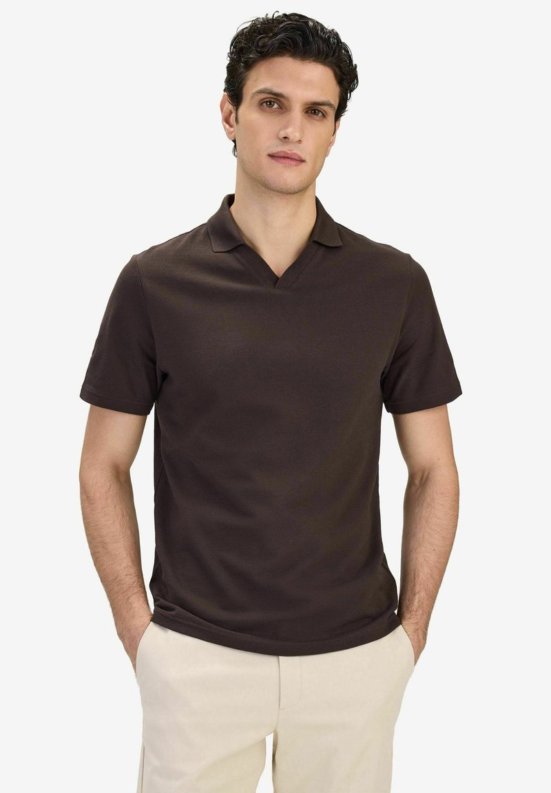 Homme aux cheveux bouclés portant un polo à manches courtes marron foncé et un pantalon beige clair, debout les mains dans les poches.