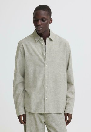 Moški, oblečen v svetlo sivo srajco z dolgimi rokavi na gumbe in ujemajoče hlače, stoji pred enostavno belo podlago.