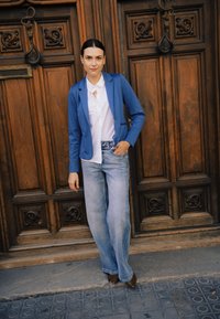 Mėlynas švarkas ant baltos palaidinės, derinamas su šviesiai mėlynomis plačiomis džinsų kelnėmis. Aprangą papildo rudi batai. Foną puošia medinės durys.