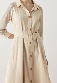 Abito camicia beige con bottoni frontali, vita cintrata e maniche in rete con un motivo a pois. Tessuto morbido con una vestibilità comoda.
