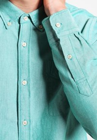 Grünblaues Hemd aus strukturiertem Stoff mit Kragen, Tasche sowie beigen Knöpfen an Vorderseite und Manschetten.