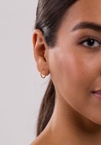 Goldene Creolen mit strukturierter Oberfläche, kreisförmigem Design und kleinen geriffelten Details. Getragen am Ohr eines Modells.