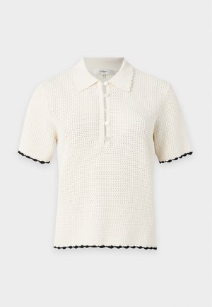 Femme debout les mains dans les poches, portant un polo en maille crème à manches courtes, un short bleu marine avec ceinture et des ballerines noires pointues sur fond blanc.