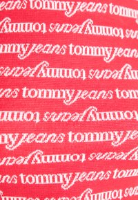 Tissu rouge présentant le texte répété "tommy jeans" dans une police cursive, mettant en valeur une texture tissée et un contraste de couleurs vif.