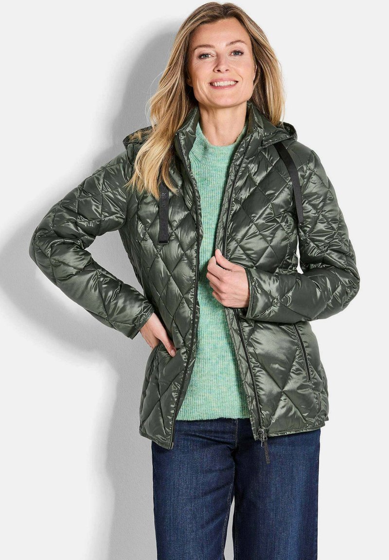 Gepolsterte olivgrüne Jacke mit Reißverschluss und Kapuze, verziert mit schwarzen Riemen und glänzender Textur, getragen über einem hellgrünen Pullover und Jeans.