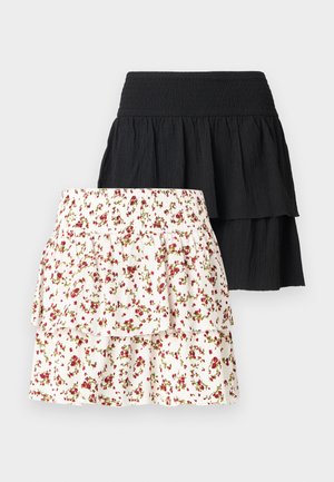 Młoda, kręcona kobieta ubrana w pomarańczowy top bez rękawów, białą kwiatową, warstwową spódnicę i brązowe sandały, stoi z ręką na biodrze.