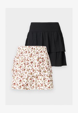 Jeune femme aux cheveux bouclés portant un haut sans manches orange, une jupe blanche à fleurs superposée et des sandales marron, debout, la main sur la hanche.