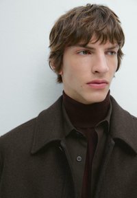 Cappotto di lana marrone con colletto, indossato sopra un dolcevita scuro e una camicia verde con bottoni. La texture è morbida; i materiali sembrano caldi.