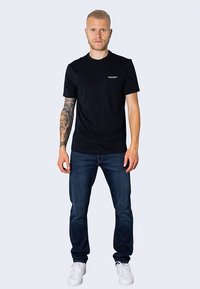 Čierne tričko s krátkym rukávom a malým bielym logom na hrudi, spárované s tmavomodrými džínsami a bielymi teniskami, stojace na neutrálnom pozadí.