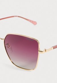 Polaroid napszemüveg aranyszínű fém kerettel, négyzet alakú lencsékkel, amelyek lila és átlátszó színátmenetet mutatnak, valamint rózsaszín díszítésű szárakkal.