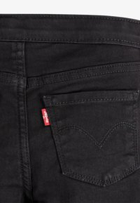 Jeans de mezclilla negra con un bolsillo trasero que presenta un patrón de costura curvada distintivo. Incluye una pequeña etiqueta de logo roja de Levi's.