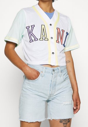 Beskåret hvid trøje med farvet tekst "KANI," mintgrønne korte ærmer, gule accenter og trykknapper; parret med lyseblå denimshorts.