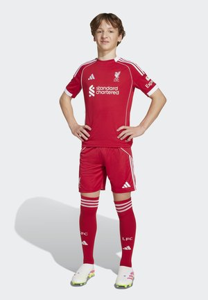 Chłopiec stoi pewnie z rękami na biodrach, ubrany w pełny czerwony strój piłkarski Liverpool FC i białe korki na białym tle.