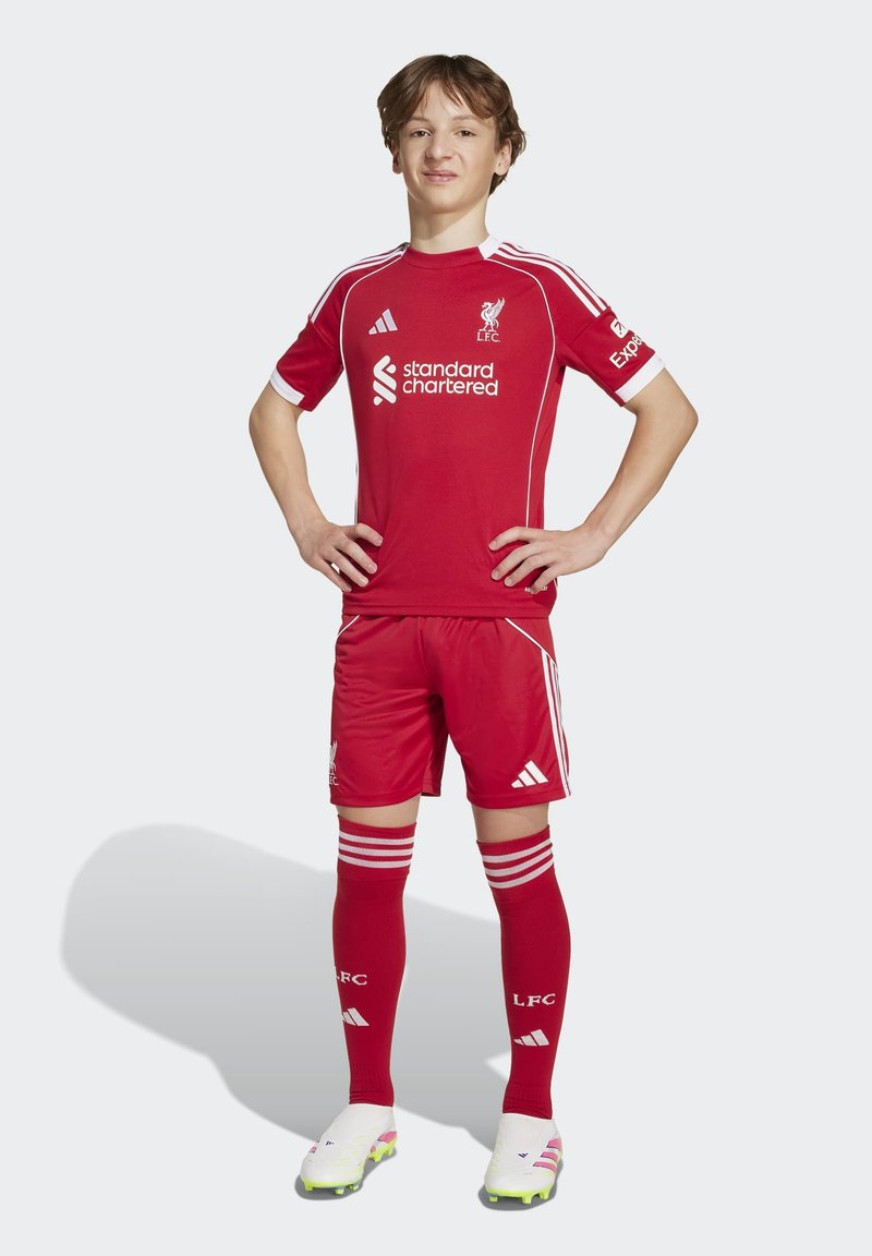 Pojke som står självsäkert med händerna på höfterna, iförd en komplett röd Liverpool FC fotbollsklädsel och vita fotbollsskor på en vit bakgrund.