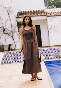 Femme en robe marron sans manches et sandales se tient à côté d'une piscine carrelée, tenant un chapeau et un carnet près d'un bâtiment blanc avec des vignes dénudées.