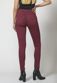 Jeans skinny burdeos con acentos de tachuelas en los bolsillos traseros y una cinturilla texturizada, combinados con botas de tobillo negras de tacón.