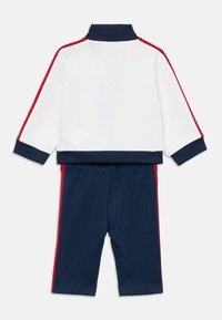 Veste de sport blanche avec des accents bleu marine et rouge, associée à un pantalon bleu marine avec des bandes latérales rouges. Col et poignets côtelés. Tissu doux.