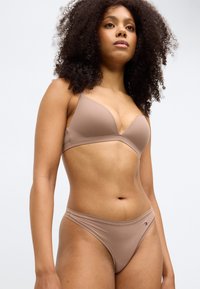 Beige-BH mit einem glatten, geformten Design und dünnen Trägern, kombiniert mit passendem Bikini-Unterteil mit minimaler Bedeckung und einem Logo-Akzent.