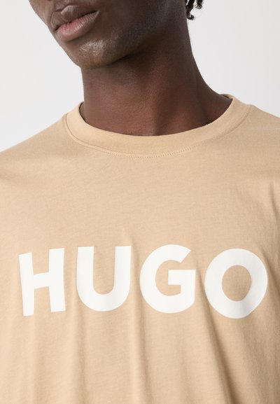 Бежова памучна тениска с кръгло деколте, с голямо бяло лого "HUGO" отпред. Мека текстура, стандартен размер.