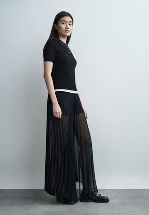 Žena v čiernom krátkom pletenom tričku s krátkymi rukávmi a dlhej priehľadnej plisovanej čiernej sukni stojaca pred jednoduchou svetlosivou stenou.