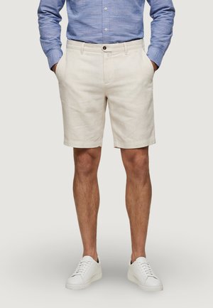 Homme portant un short beige, une chemise bleue à manches longues boutonnée et des baskets blanches, debout les mains dans les poches sur un fond neutre.
