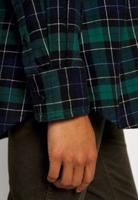 Chemise en flanelle à motif à carreaux noirs et verts avec des accents blancs. Détail du poignet boutonné visible, avec des boutons foncés contrastant avec la texture du tissu.